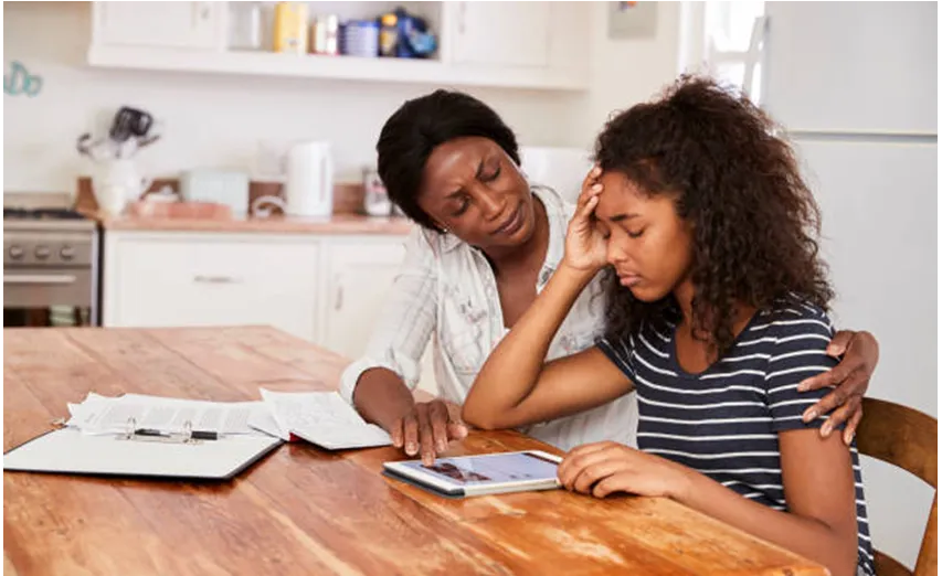A mother comforting her teenage daughter as she works through exam papers at the kitchen table